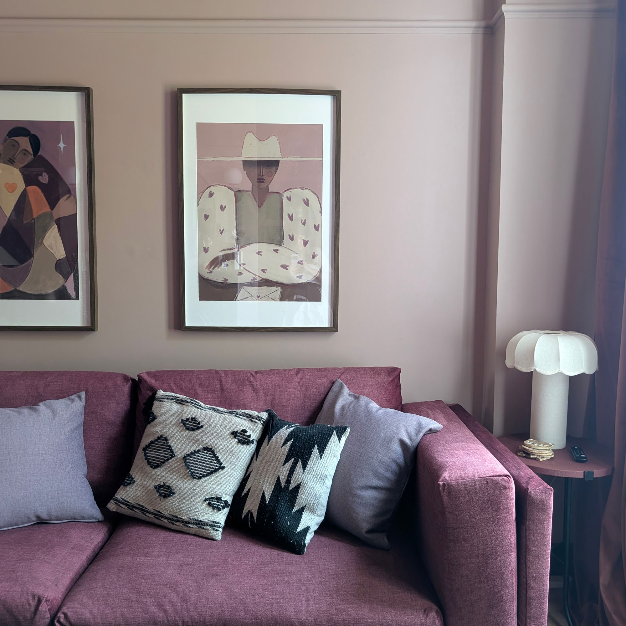 Pale pink living room with framed paintings of cowgirls on wall above dark pink sofa with black and white patterned and solid purple coloured cushions
