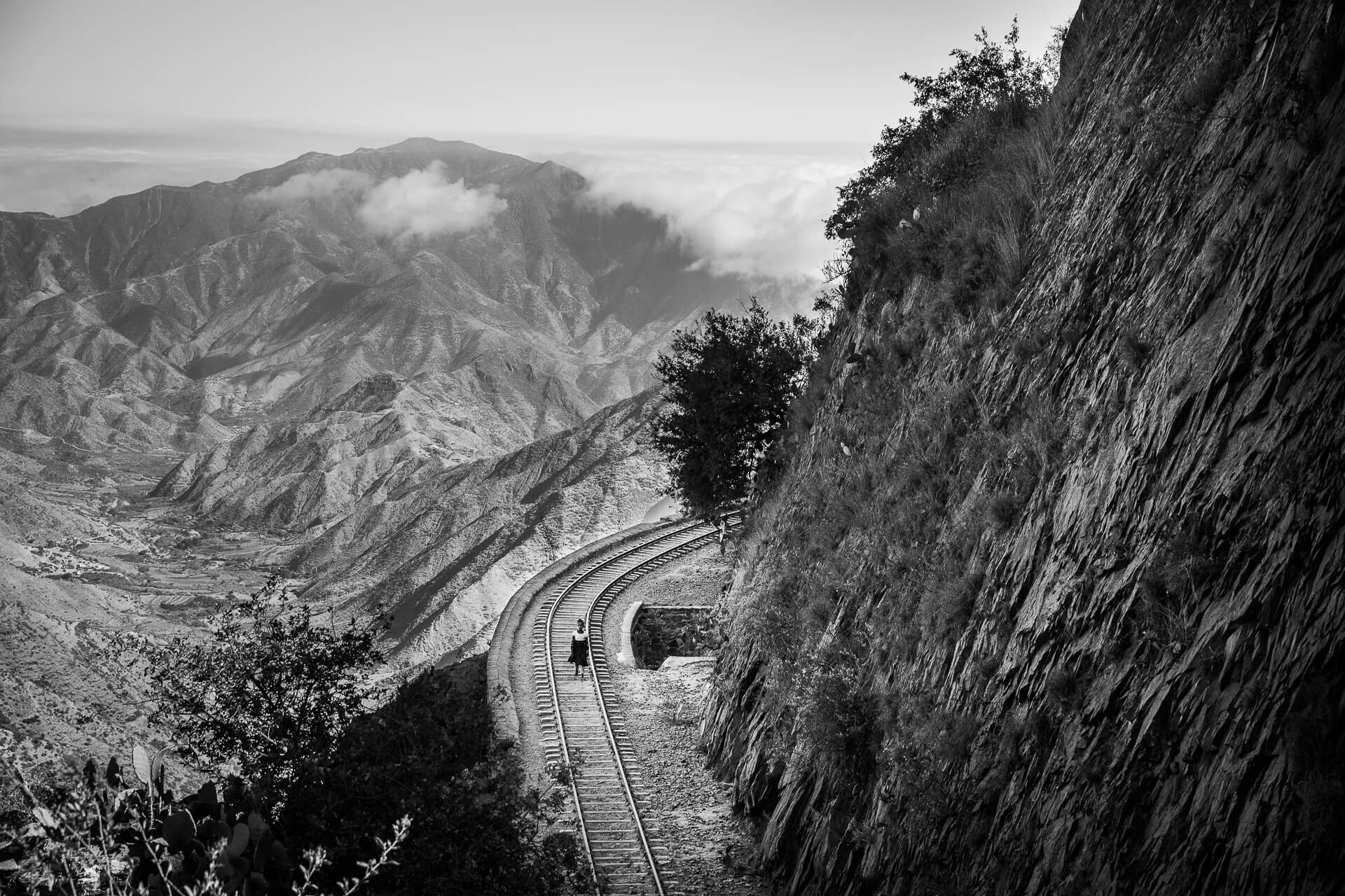 Monochrome image of women walking along mountainside rail line.