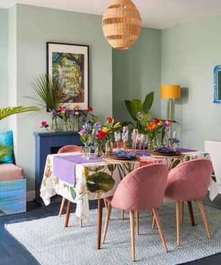 A mint green dining room with a colorful dining room with two vases of flowers and a purple dining runner, dusky pink velvet chairs, and a white rug underneath