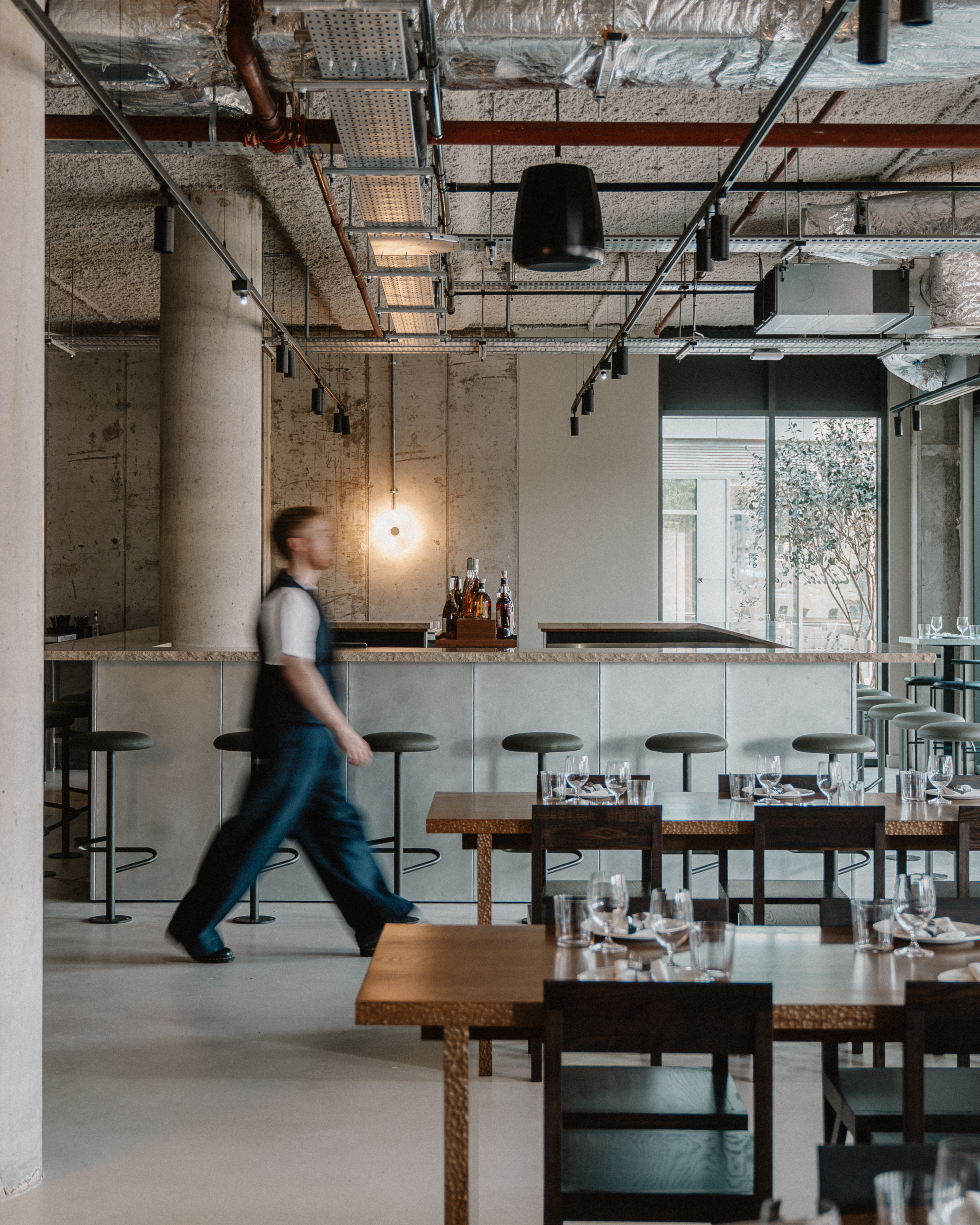 A young man dressed in navy blue trousers and vest and a white T-shirt is captured in movement as he walks through the dining room of a modern restaurant with brutalist cement, chrome, and wood interiors.