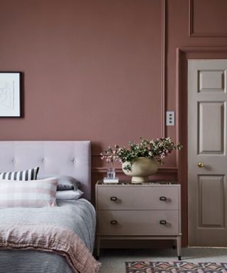 A light, dusty pink bedroom with a bed with padded headboard, wide bedside table and a green plant