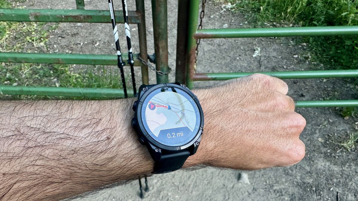 The Garmin Fenix 8 showing a course map with the location "off trail" by 0.2 miles in front of a gate, with a pair of trekking poles leaning against the gate.