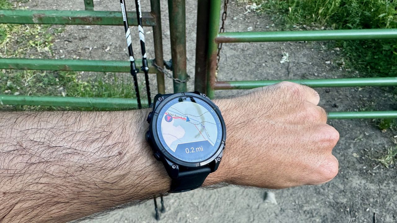 The Garmin Fenix 8 showing a course map with the location "off trail" by 0.2 miles in front of a gate, with a pair of trekking poles leaning against the gate.