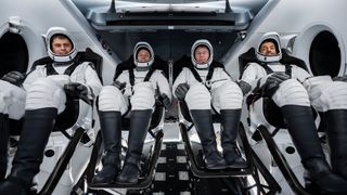 four spacesuited astronauts sitting side by side in a spacecraft with visors up and seatbelts on
