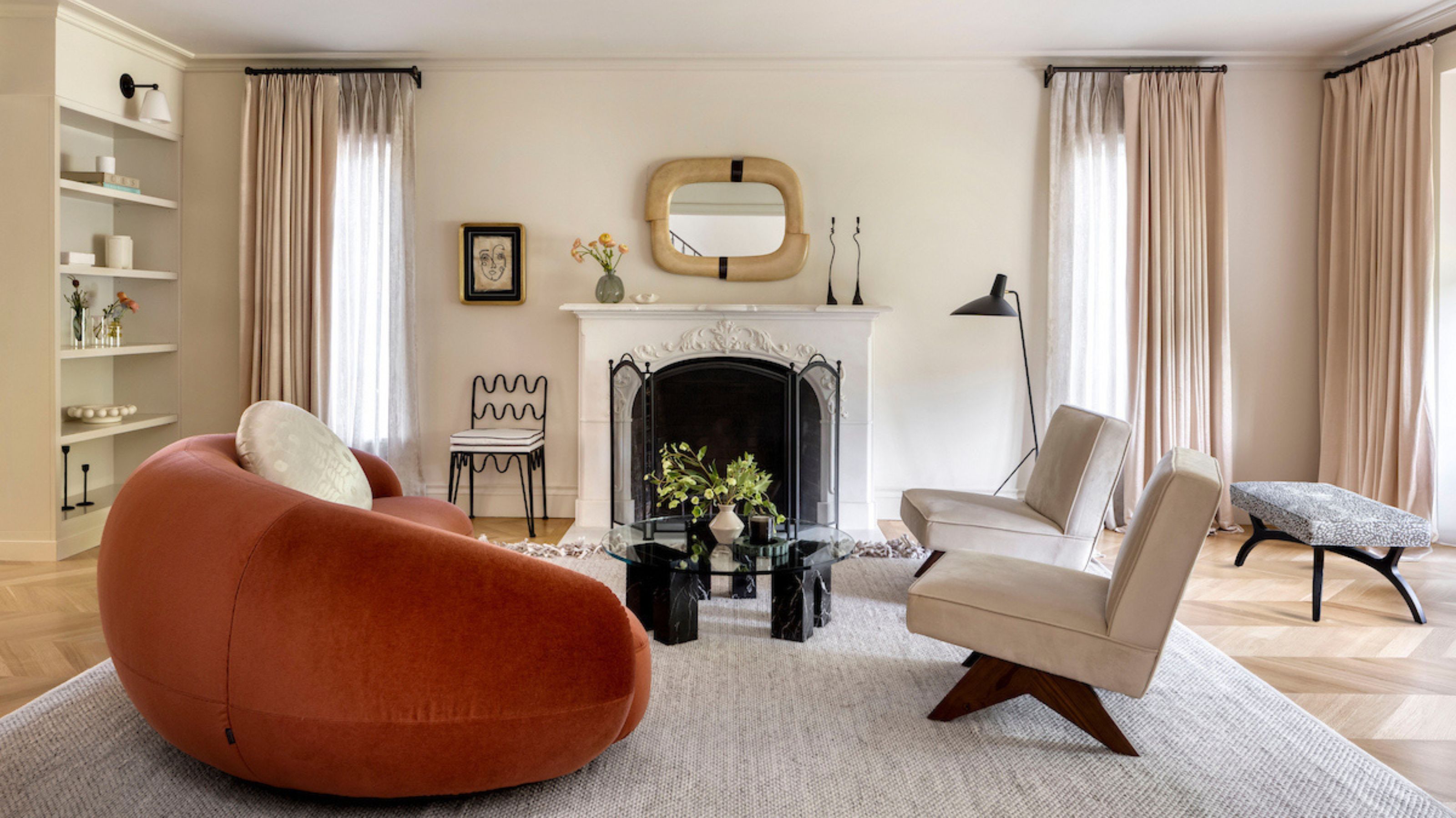 A living room with an orange/red sofa and two cream colored accent chairs opposite it. There is also a fireplace close by with a mirror and accessories above it. On one side of the living room is open shelving with decor pieces placed inside