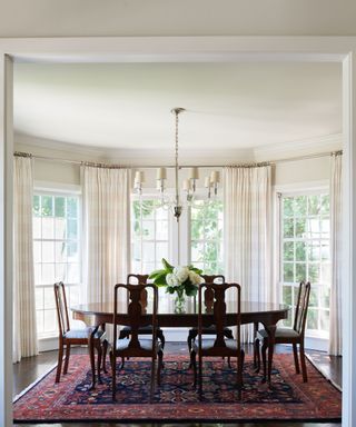 Old money coastal dining room with a vintage dark wood dining table and chairs, a red and dark blue Persian rug, and contemporary light and curtains