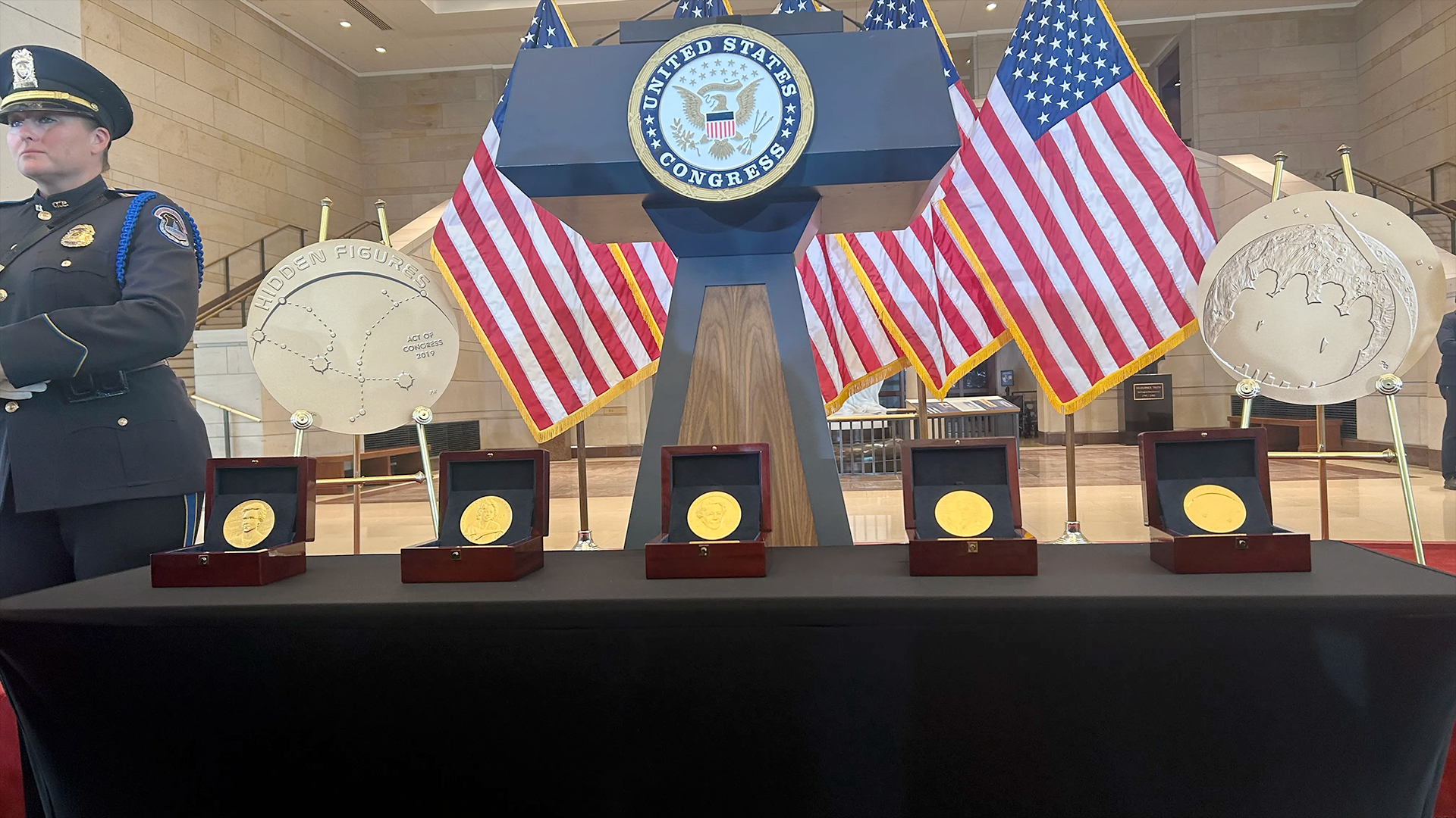 closeup photo of five gold medals arrayed on a stage in front of a lectern with the presidential seal on it, with american flags in the background