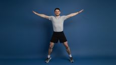 Smiling man in shorts and T-shirt with his arms outstretched and legs apart, mid star jump, against a blue background