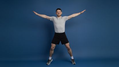Smiling man in shorts and T-shirt with his arms outstretched and legs apart, mid star jump, against a blue background