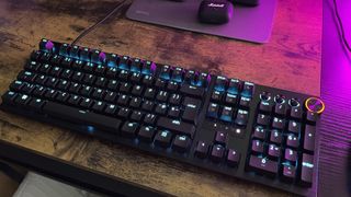 The Razer Huntsman V3 Pro is shown pictured on a wood effect desktop with a purple light coming from the right of the image. The keys are illuminated in pale blue.