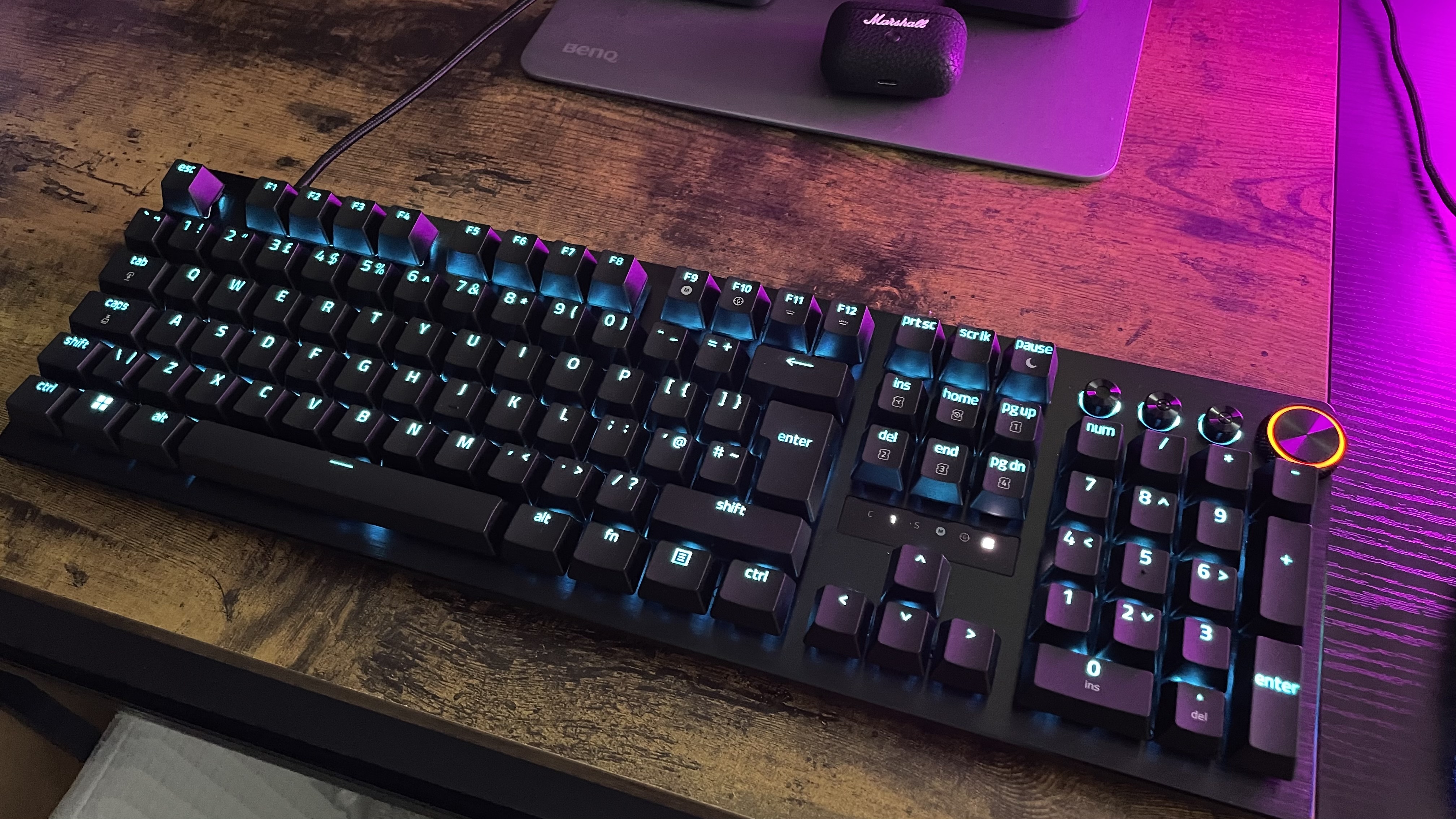 The Razer Huntsman V3 Pro is shown pictured on a wood effect desktop with a purple light coming from the right of the image. The keys are illuminated in pale blue.
