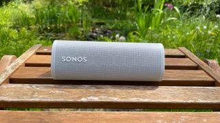 A white Sonos Roam speaker on its side on a wooden slatted deckchair in front of some plants.