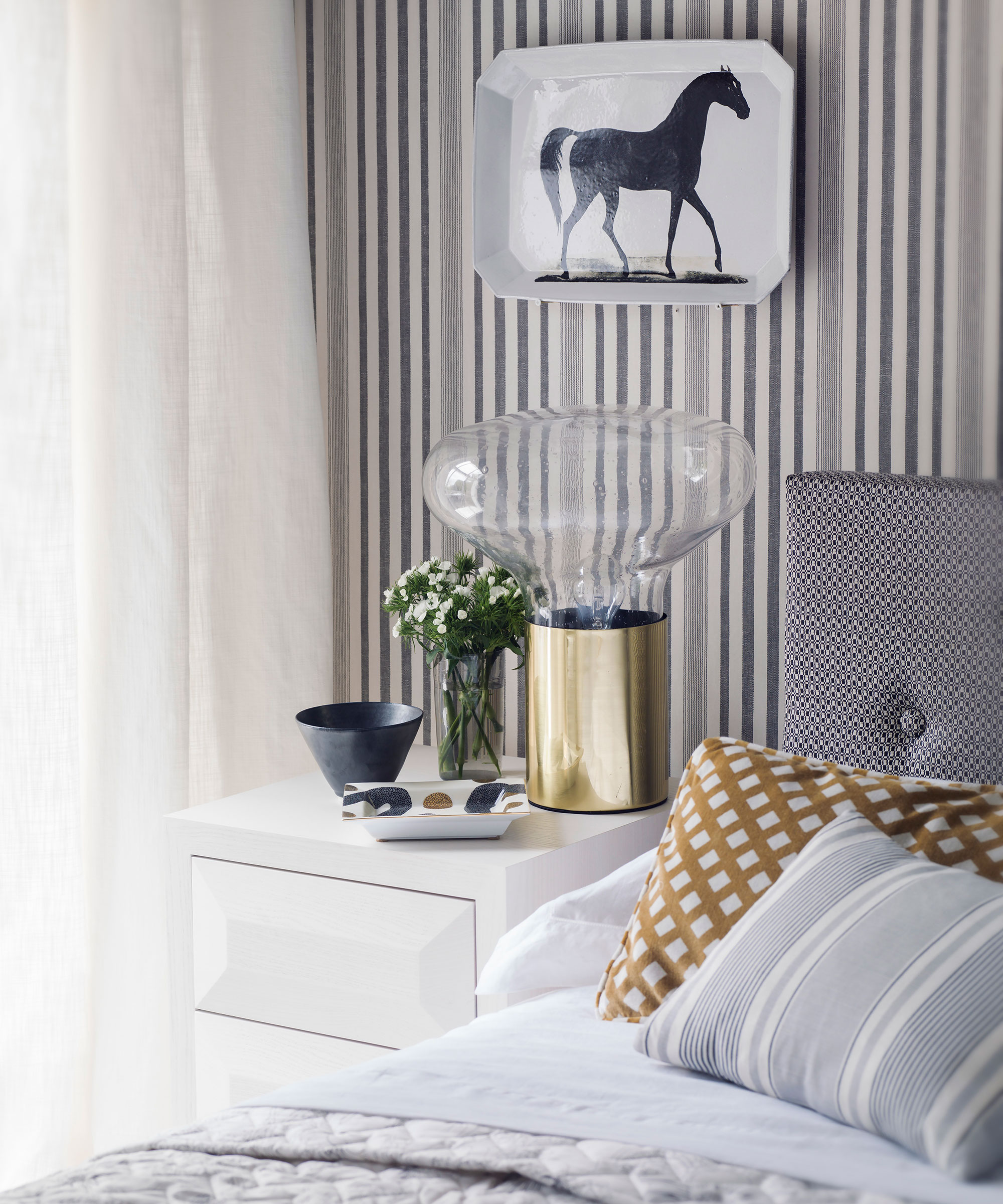 An example of bedroom lighting ideas showing an oversized glass and gold table lamp in front of striped wallpaper
