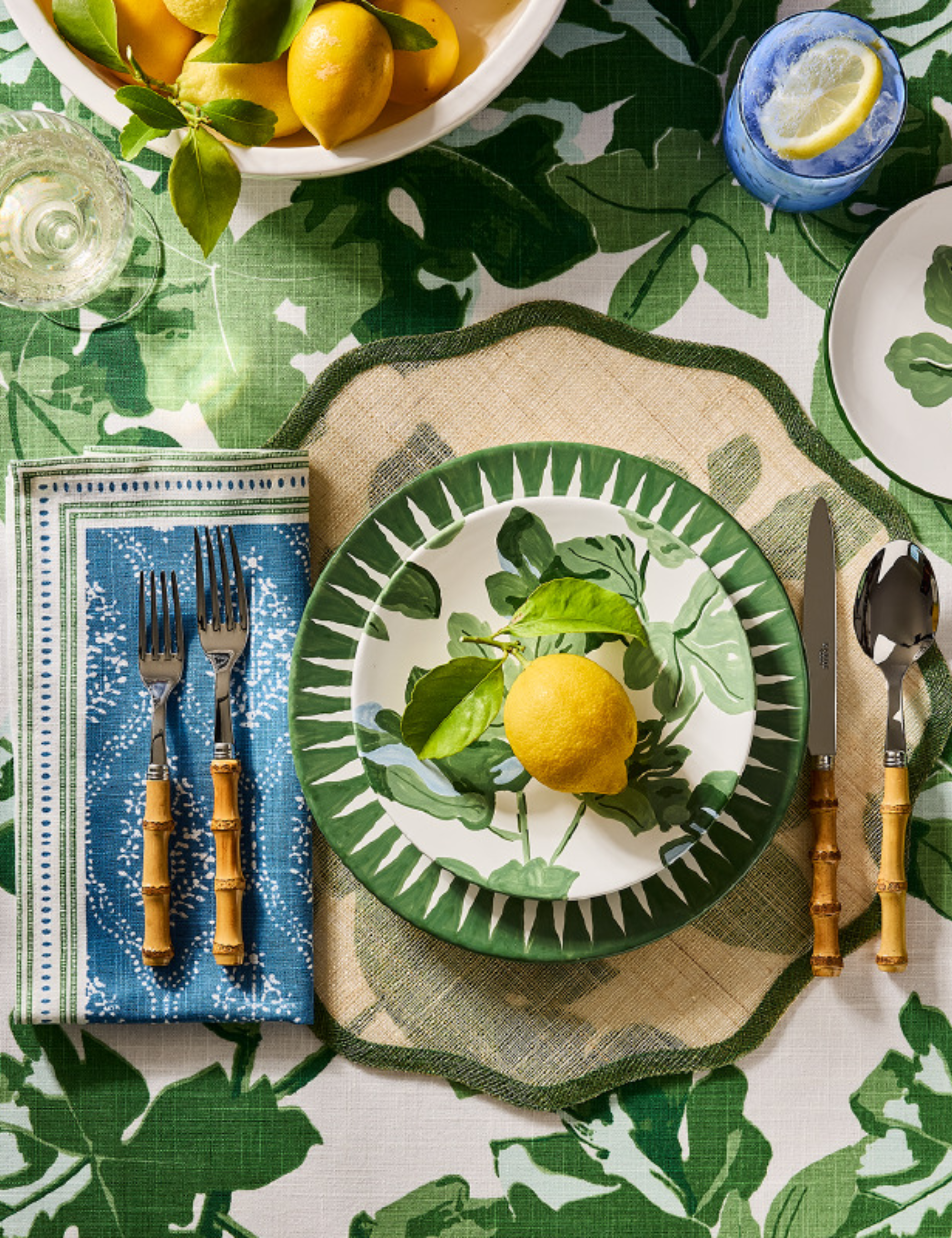 Green fig patterned tablescape featuring a scalloped placemat, stacked verdant green plates, bamboo handled cutlery, and blue linen napkins.