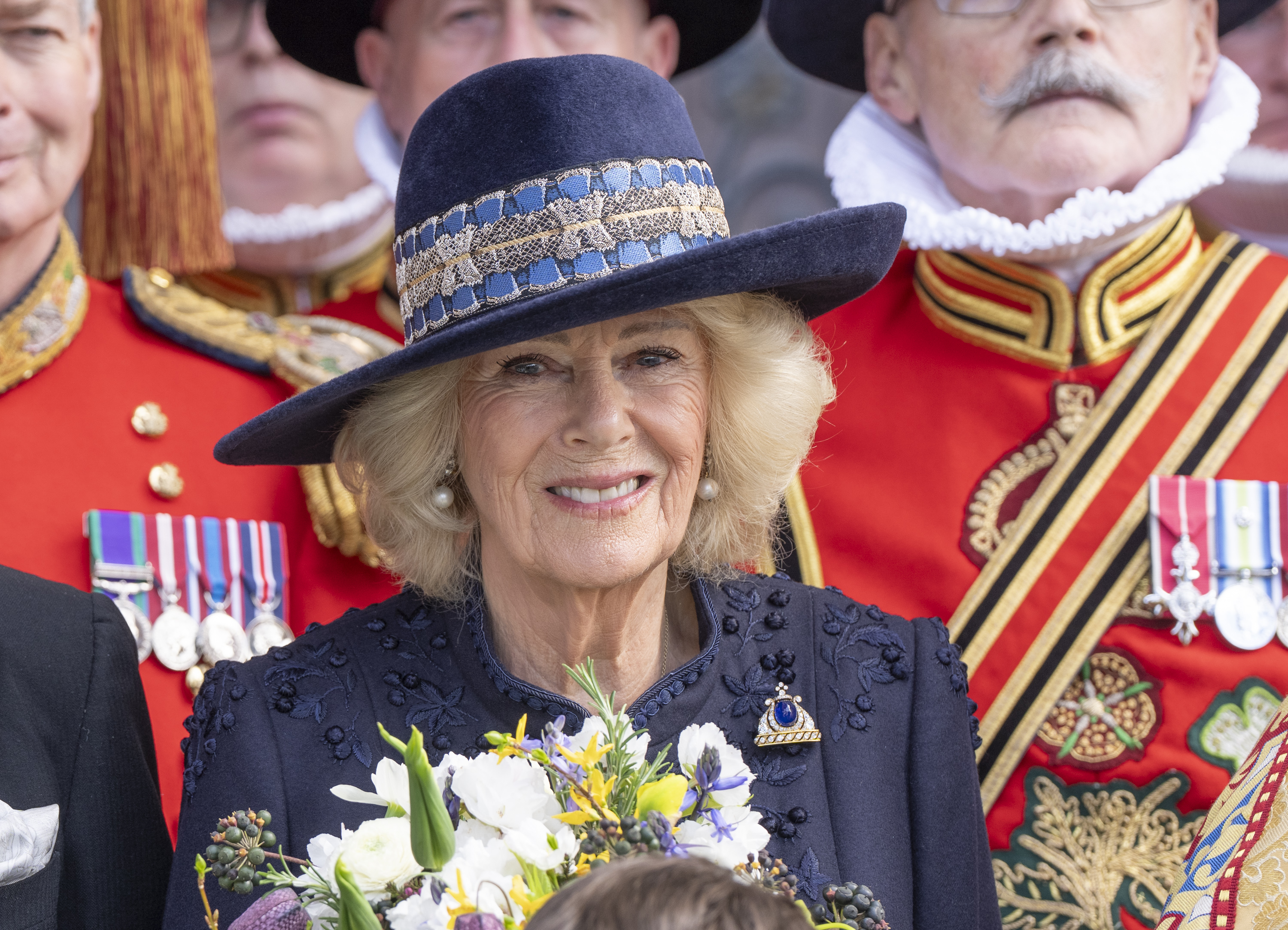Queen Camilla wearing a blue coat and hat standing in front of beefeaters