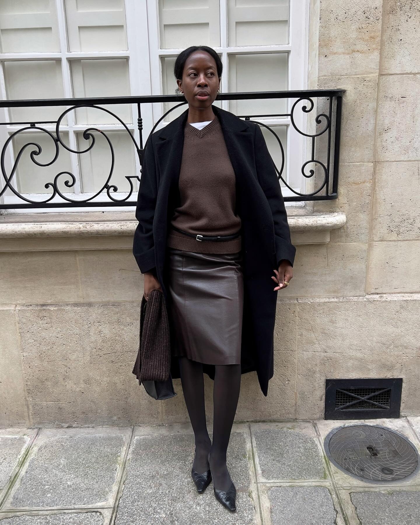 Sylvie wears a black wool coat, brown v-neck sweater, brown belt, brown leather skirt, brown tights, and black slingbacks.
