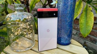 En Google Pixel 6 står utomhus på ett litet träbord, med sitt unika färgschema som skiftar i vitt, svart och rött på baksidan av mobilen.
