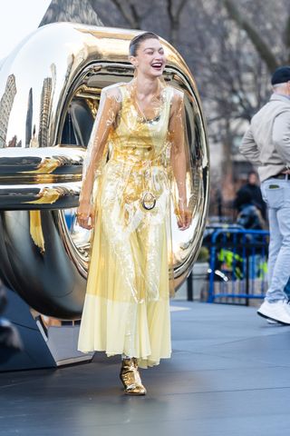 A photo of Gigi Hadid sporting a sheer gold trench coat over a butter yellow sleeveless dress.