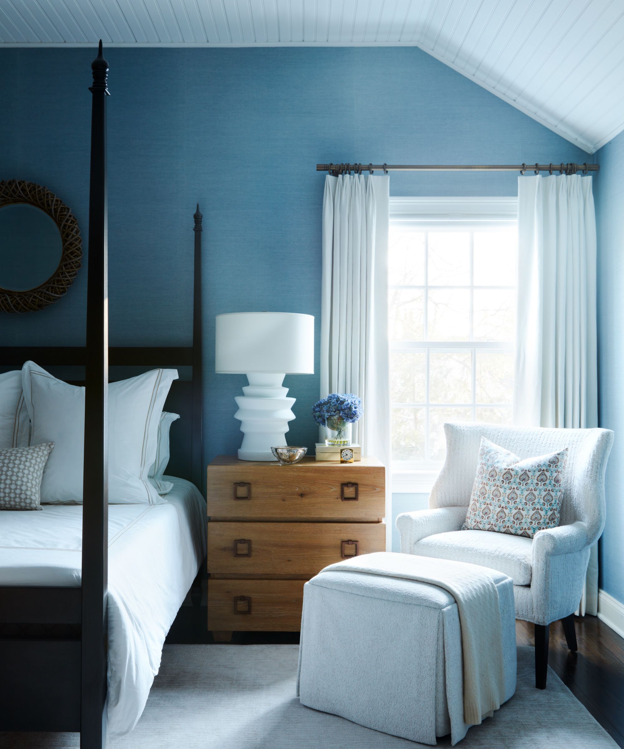 Bedroom with blue painted walls, a dark wood mahogany four-poster bed, white bedding, a wooden nightstand, and a cream armchair with a cream linen footstool.