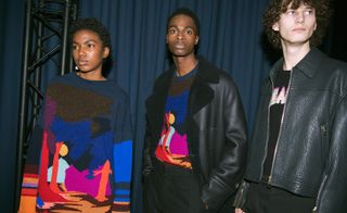 Three models wearing looks from Paul Smith's collection. Two models are wearing jumpers with a multicoloured design and one of them is wearing black trousers and black coat with short fur collar. The third model is wearing a black top with multicoloured wording, black trousers and a dark green textured leather jacket