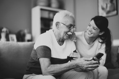 Senior adult father and his adult daughter smiling, sitting on a sofa, looking at a mobile phone together