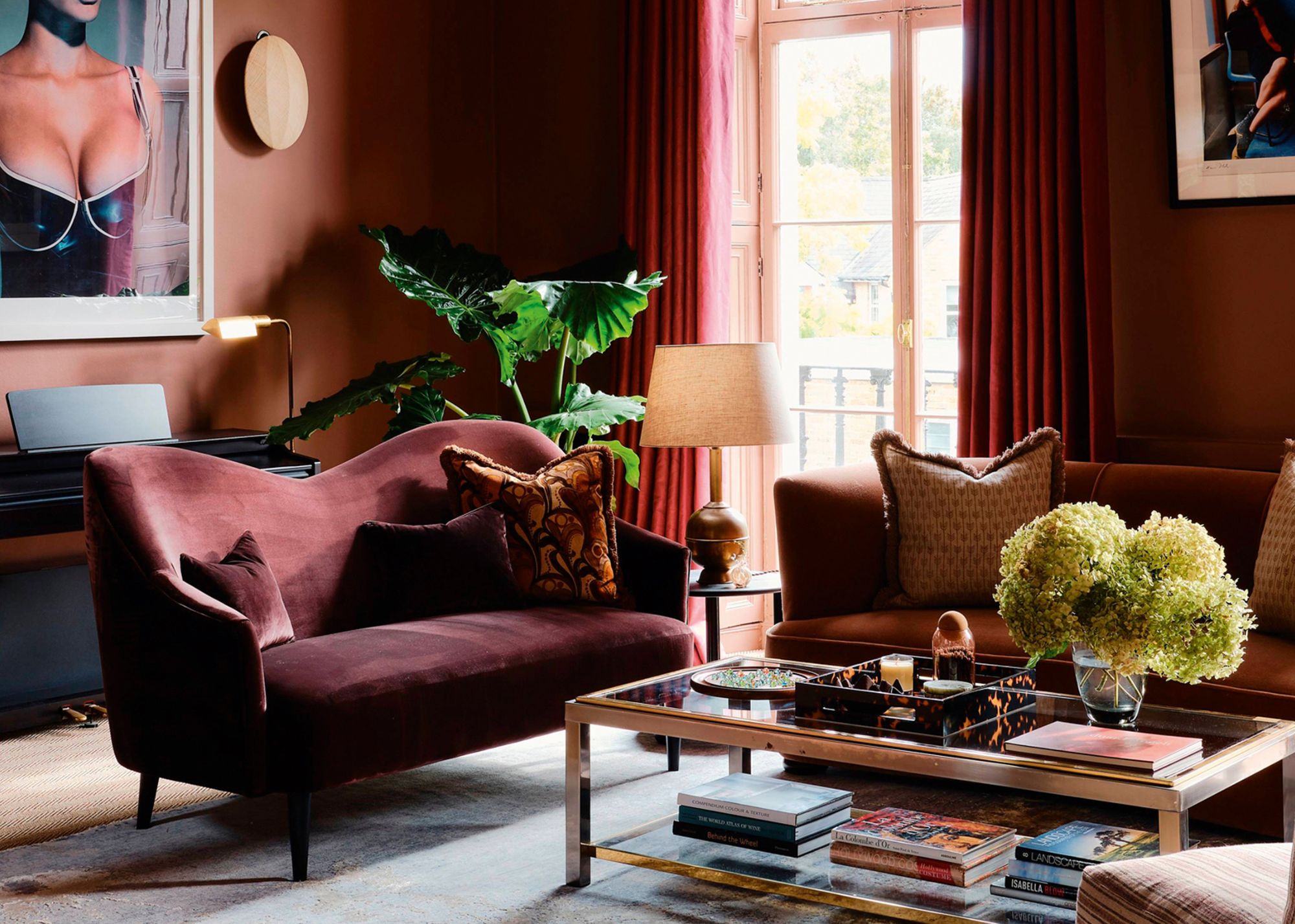 A modern living room decorated with bold earth tones with red curtains, a burgundy sofa next to a brown sofa in front of a rectangular glass coffee table with flowers and decor on top
