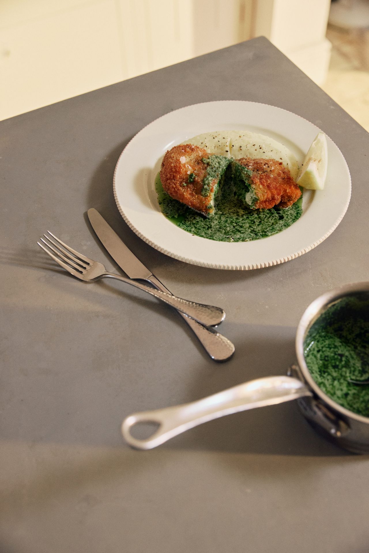 The corner of a metal countertop with a white plate that has food on it and a knife and fork beside it.