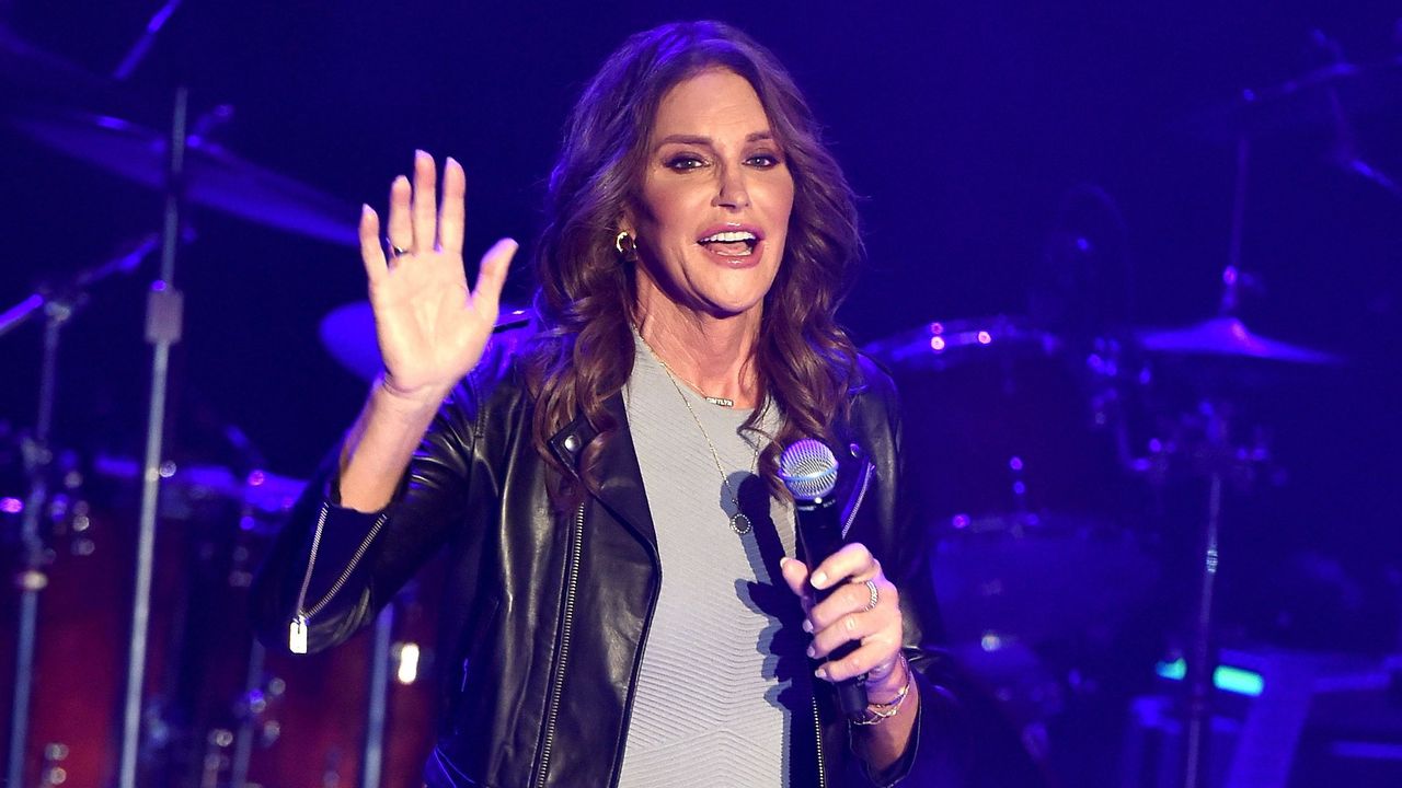 Caitlyn Jenner waving, wearing a black leather jacket holding a mic