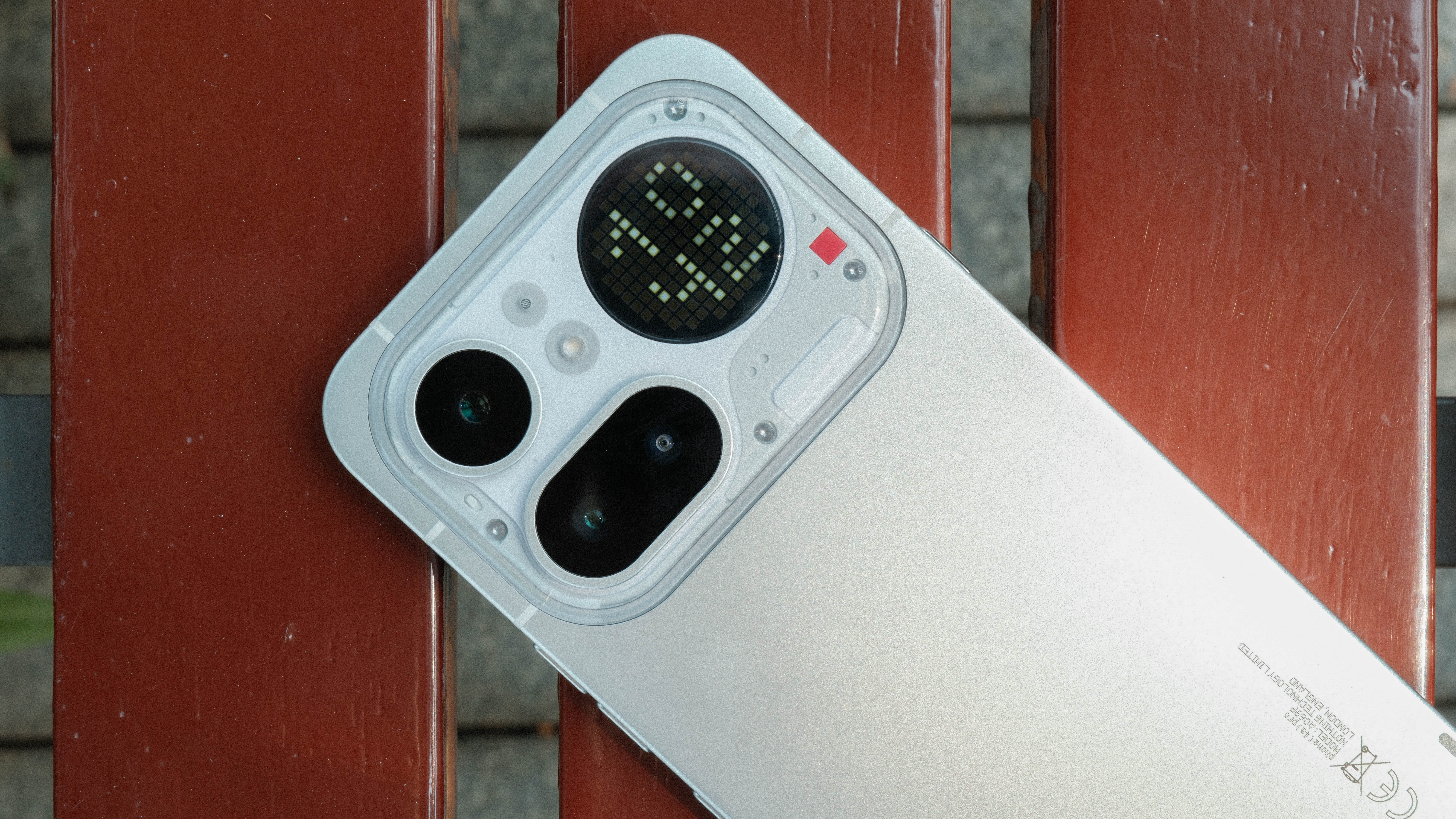 A top-down view of a light gray smartphone's camera module. The module is encased in a transparent raised border, containing two large black camera lenses, two smaller sensors, a small red square accent, and a circular LED dot-matrix display.