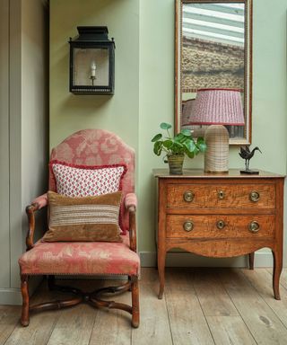 A red armchair with two scatter cushions on it. Above the chair is a lantern wall light and next to the chair is a chest of drawers with a plant, a sculpture and a lamp on it. The lamp has a red lampshade. The walls are painted a mint green colour.