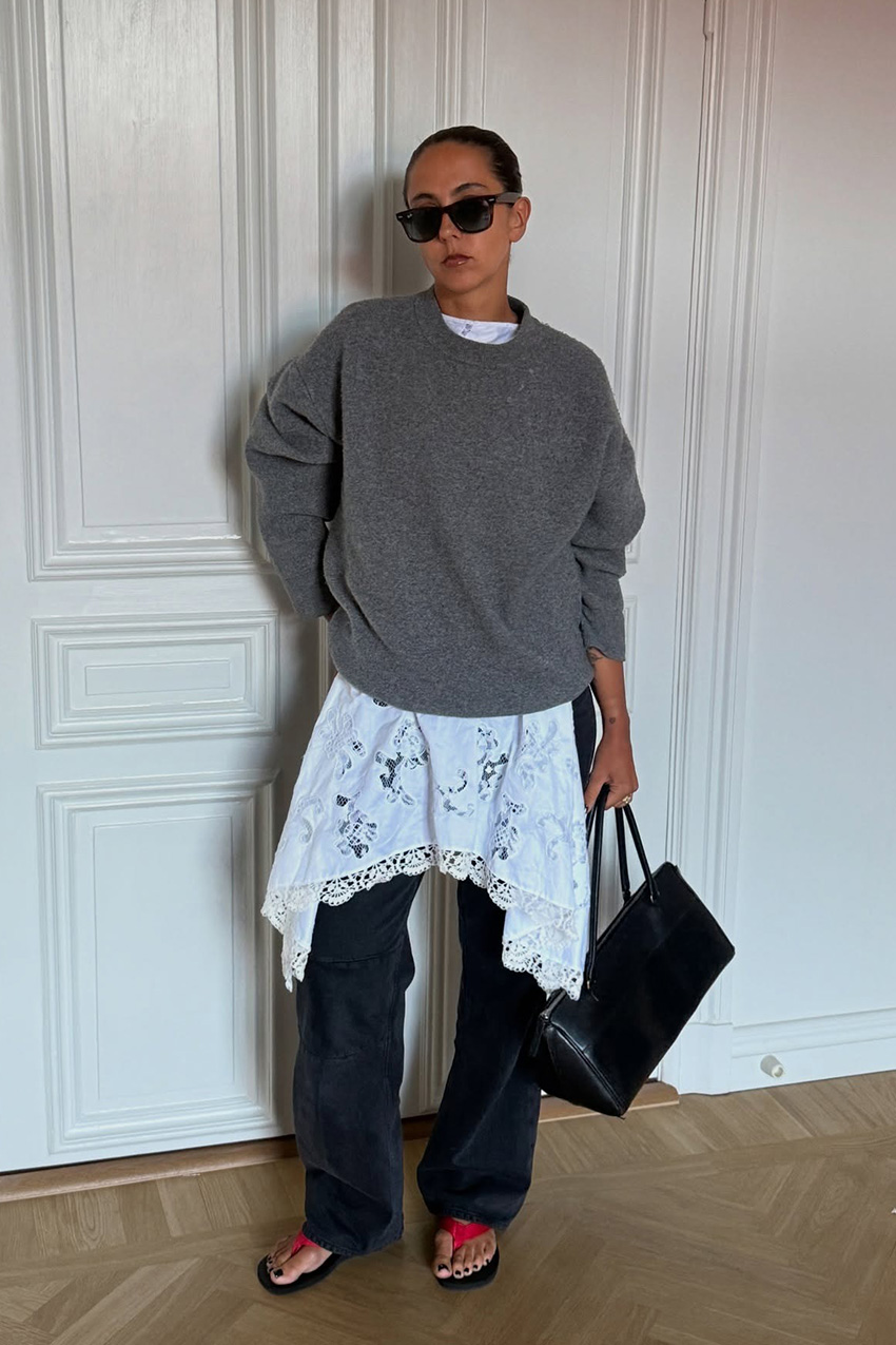 A sweater styling trend is shown in a photo of a woman&#039;s outfit, where she&#039;s standing inside her home against a white wall, wearing an oversize gray crewneck sweater over a white lace slip dress styled over black baggy jeans with red and black flip flops, black sunglasses, and a black handbag