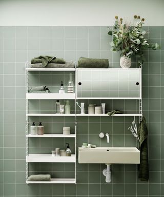 Green bathroom with green tiles, showing modular string shelving unit in white, shelf, cabinet, mirror and hook sections, shelf positioned above small white sink
