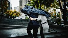 A couple run arm in arm up steps while ducking under a jacket in the rain.