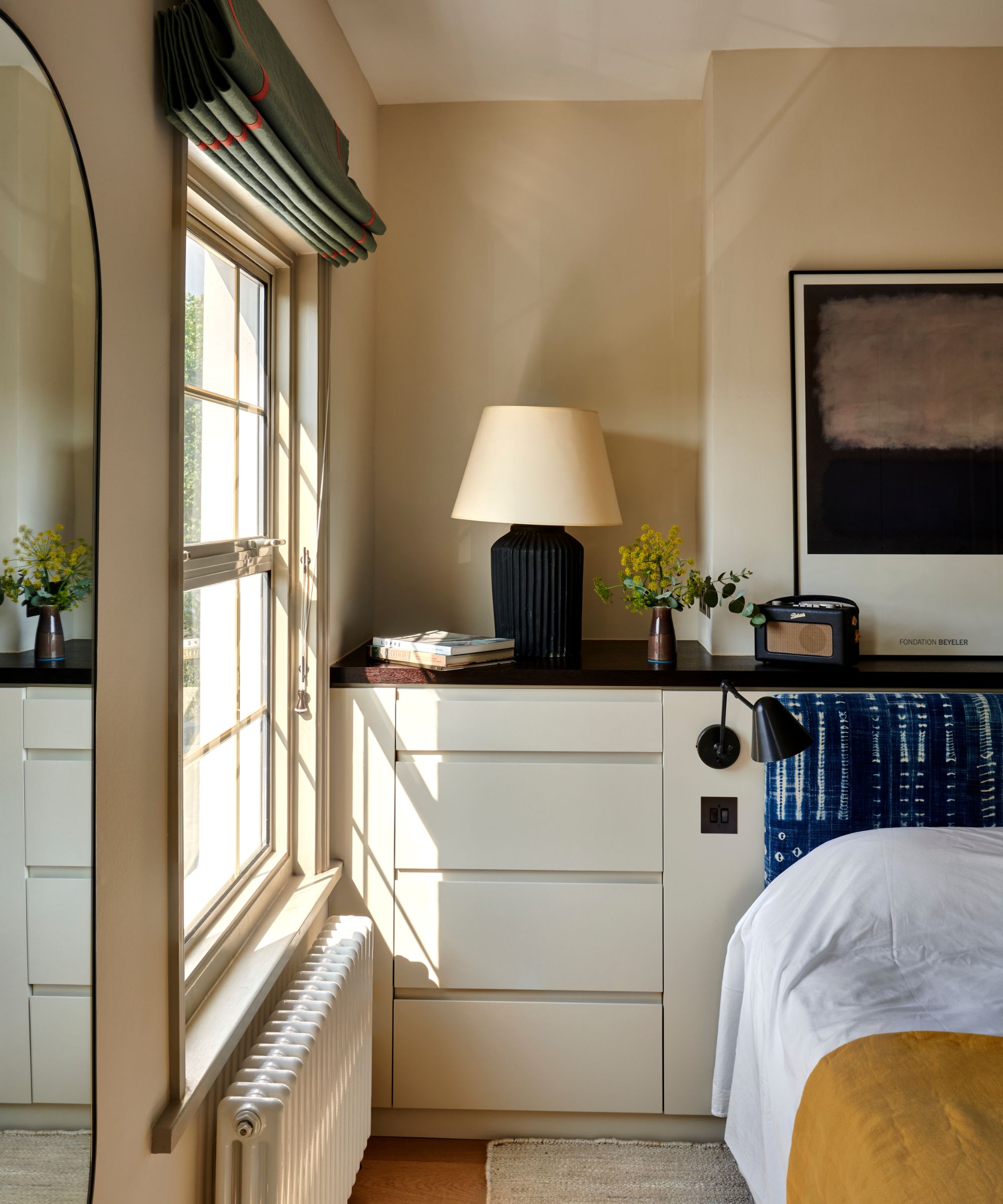 Bedroom with drop cloth by Farrow and Ball on the walls, large window with green roman blind, built-in drawers, a blue indigo headboard and white and yellow bedding