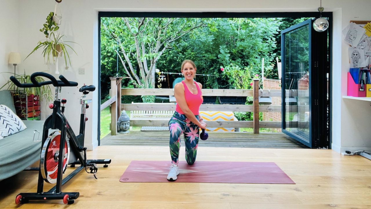 Personal trainer Maddy Biddulph performs a kneeling woodchop. She wears a pink vest, tropical print leggings and white sneakers. She is half-kneeling, with her left knee on the floor and her right leg at a 90 degree angle in front of her. She holds a kettlebell in both hands close to her left hip. Behind her is a leafy garden and next to her is an exercise bike.