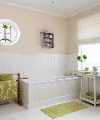 A bathroom with white shiplap and a beige wall, a stained glass circular window, a gray wall shelf, and a rectangular window, an off-white two-tier shelf, a bath, and a wooden towel rail with green towels