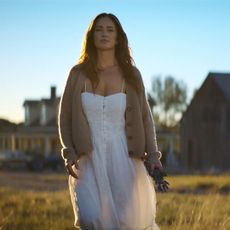 Minka Kelly as Quinn, wearing a white dress and cardigan, holds flowers as she walks through a field with a farmhouse behind her, in Episode 202 of Netflix's 'Ransom Canyon.'