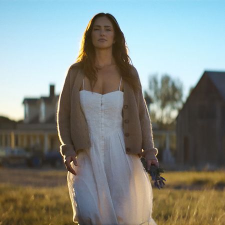 Minka Kelly as Quinn, wearing a white dress and cardigan, holds flowers as she walks through a field with a farmhouse behind her, in Episode 202 of Netflix's 'Ransom Canyon.'
