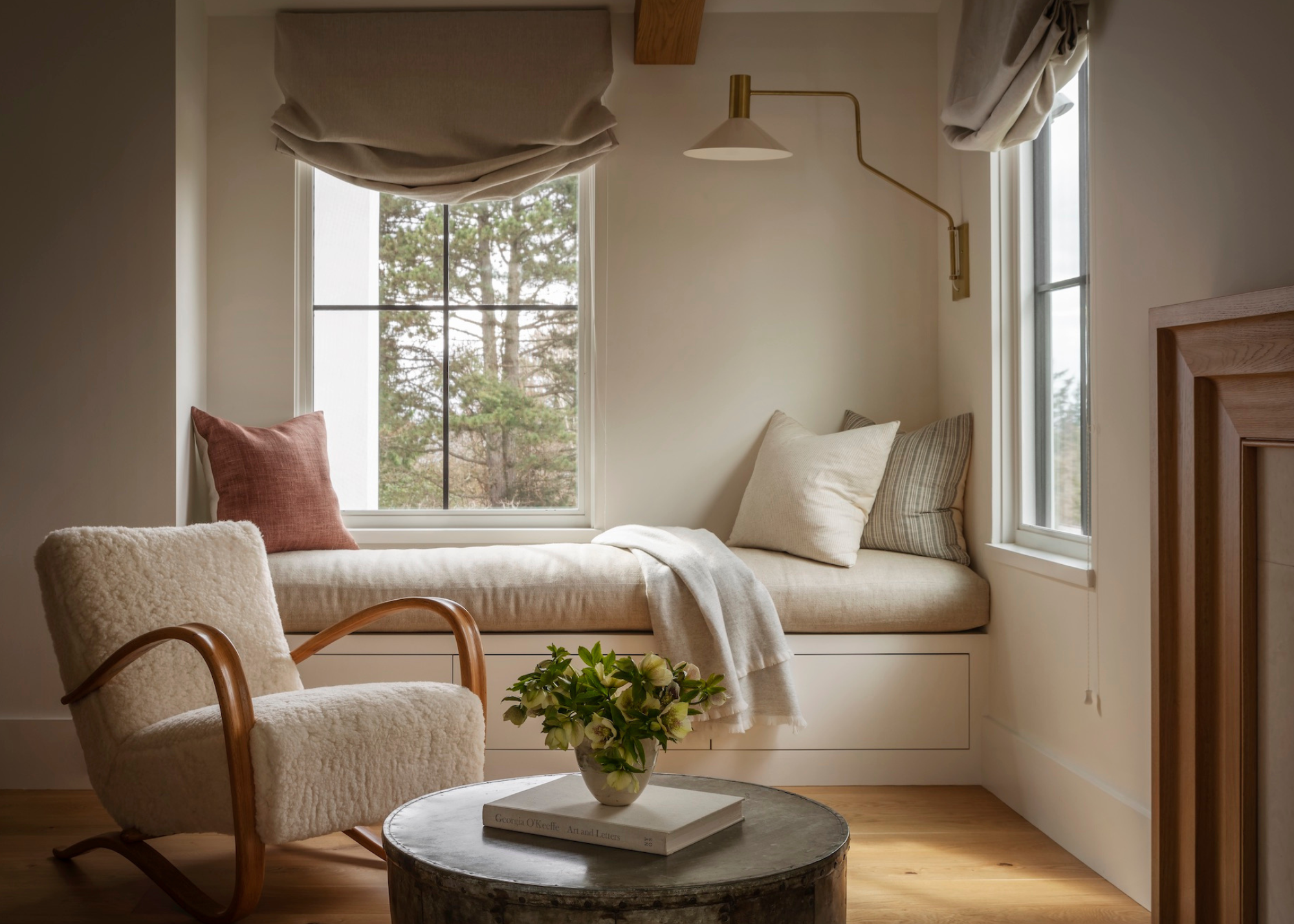 a living room with built-in window seat with storage in a scandi cool aesthetic with blinds a modern wall light and boucle accent chair next to a coffee table with flowers on it next to the fireplace