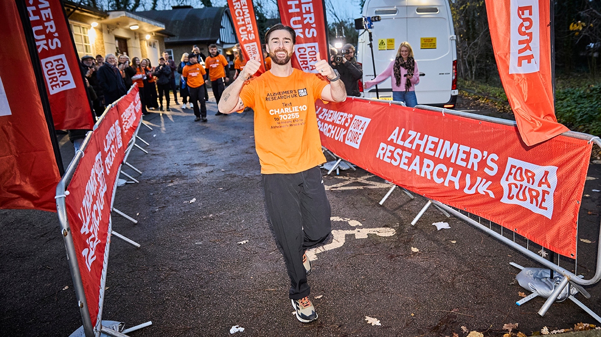 Charlie Quirke smiling, in an orange t-shirt and black tracksuit bottoms with his thumbs up. In the background are orange banners for Alzheimer's Research UK, Charlie's supporters and a cameraman.