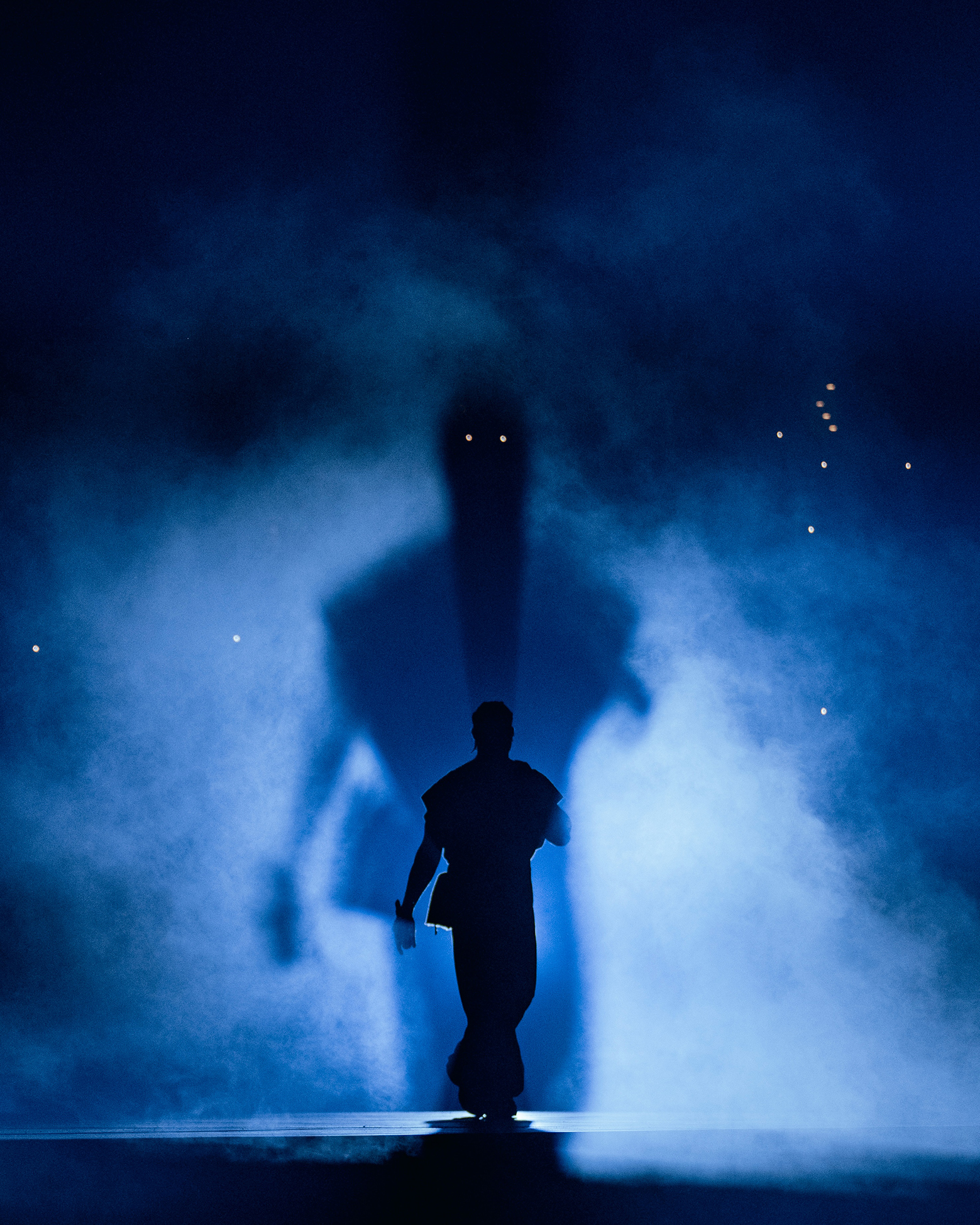 Artist Drake standing on stage, blue background, surrounded by smoke, showcasing his shadow behind him