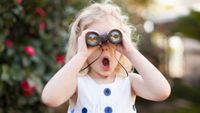Little girl has a shocked expression while using one of the best binoculars for kids with a foliage backdrop