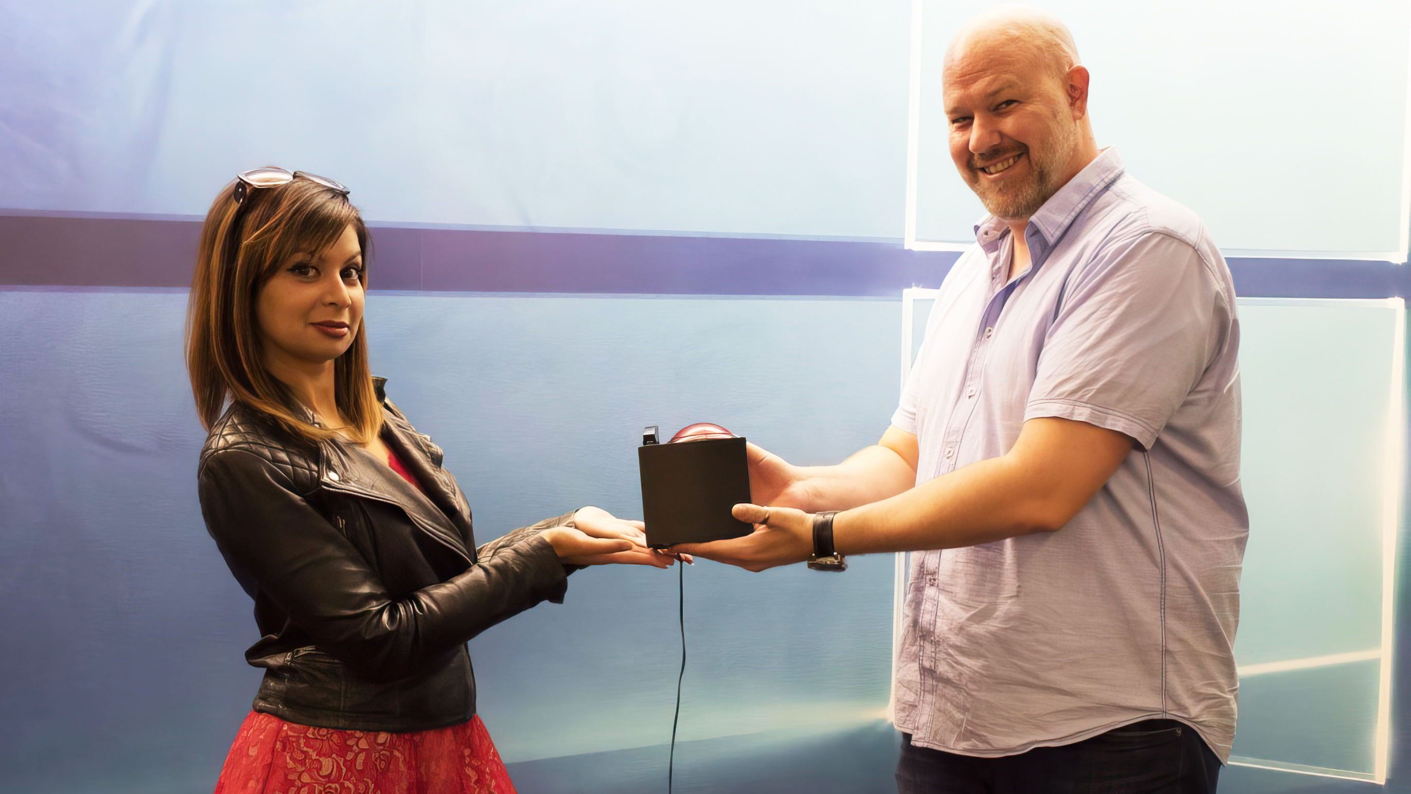 A woman in a black leather jacket passes a small black box to a smiling man in a light shirt. They are standing in front of a blue background.