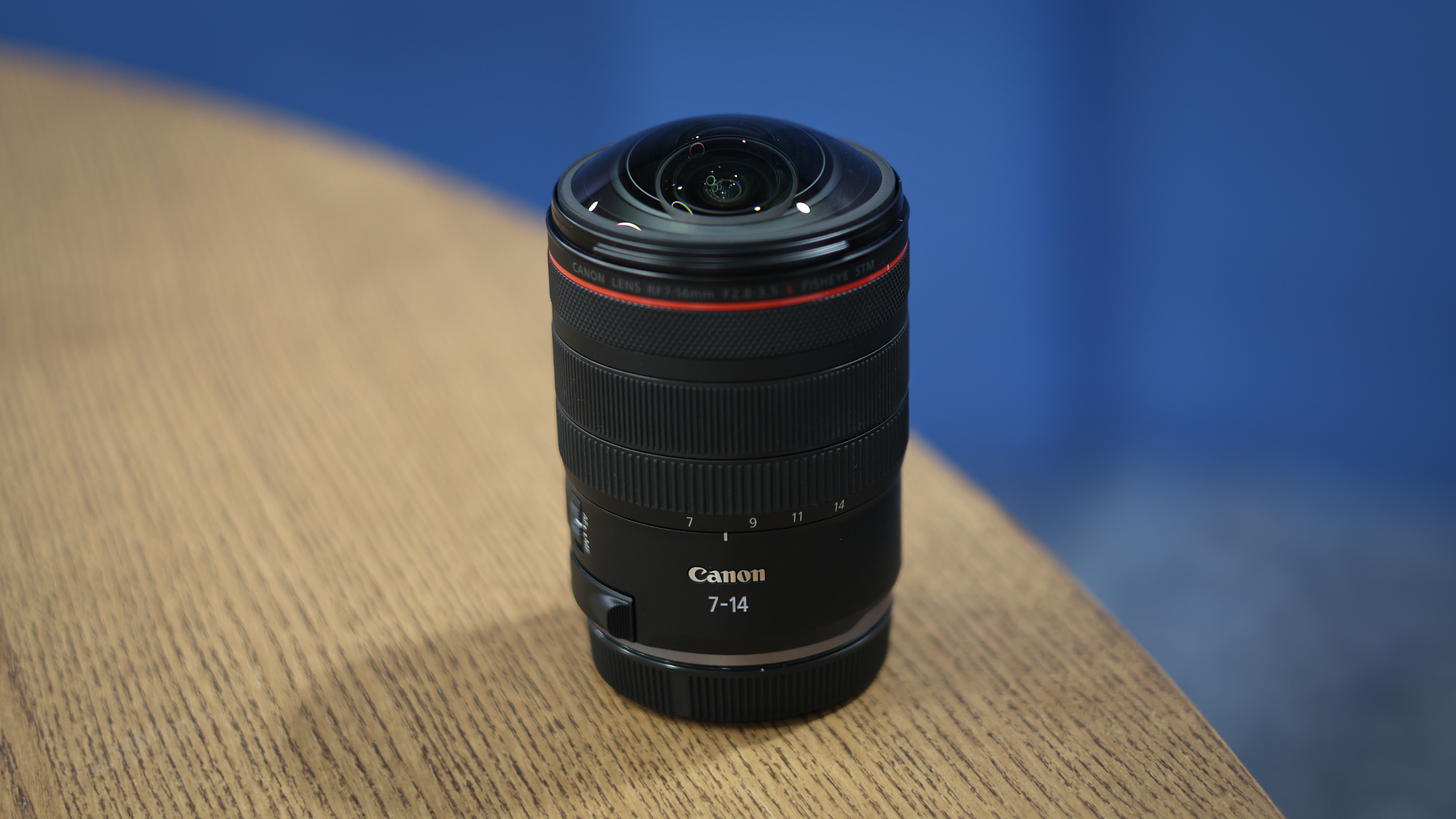 Canon RF 7-14mm f/2.8-3.5L Fisheye STM on a wooden surface in front of a blue background