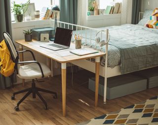 A cute, modern college dorm room with a wooden desk placed at the tip of the bed with a blue comforter and multicolored pillow next to a window with plants