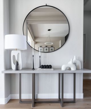 Round black trim mirror above a console table staged with candle holders, decorative pots and a white table lamp