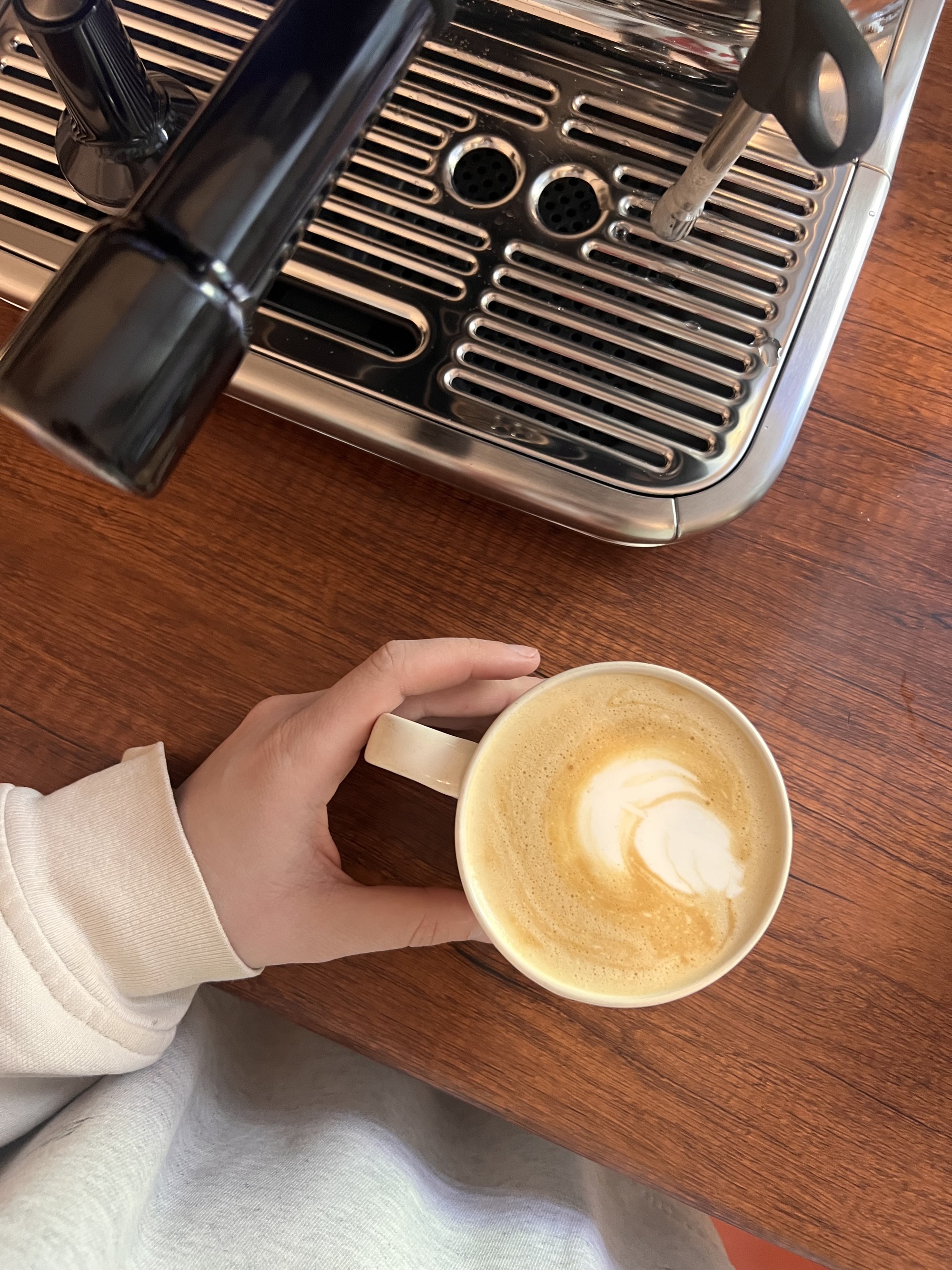 Hand holding a coffee mug with a latte in it on a wooden countertop with a silver coffee machine beside it.