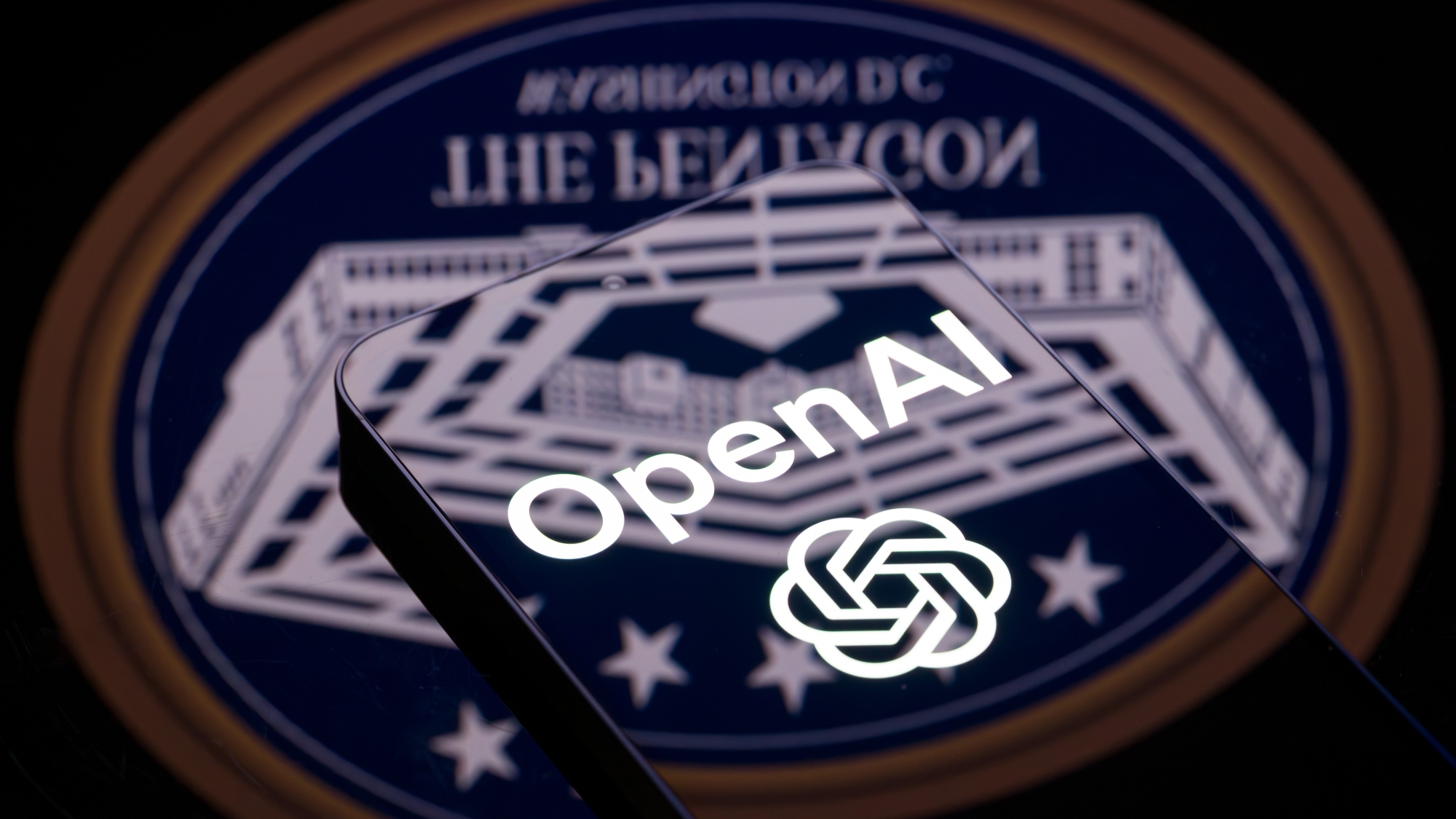 The OpenAI logo is displayed on a smartphone with the seal of the U.S. Department of Defense (Pentagon) reflected on a surface
