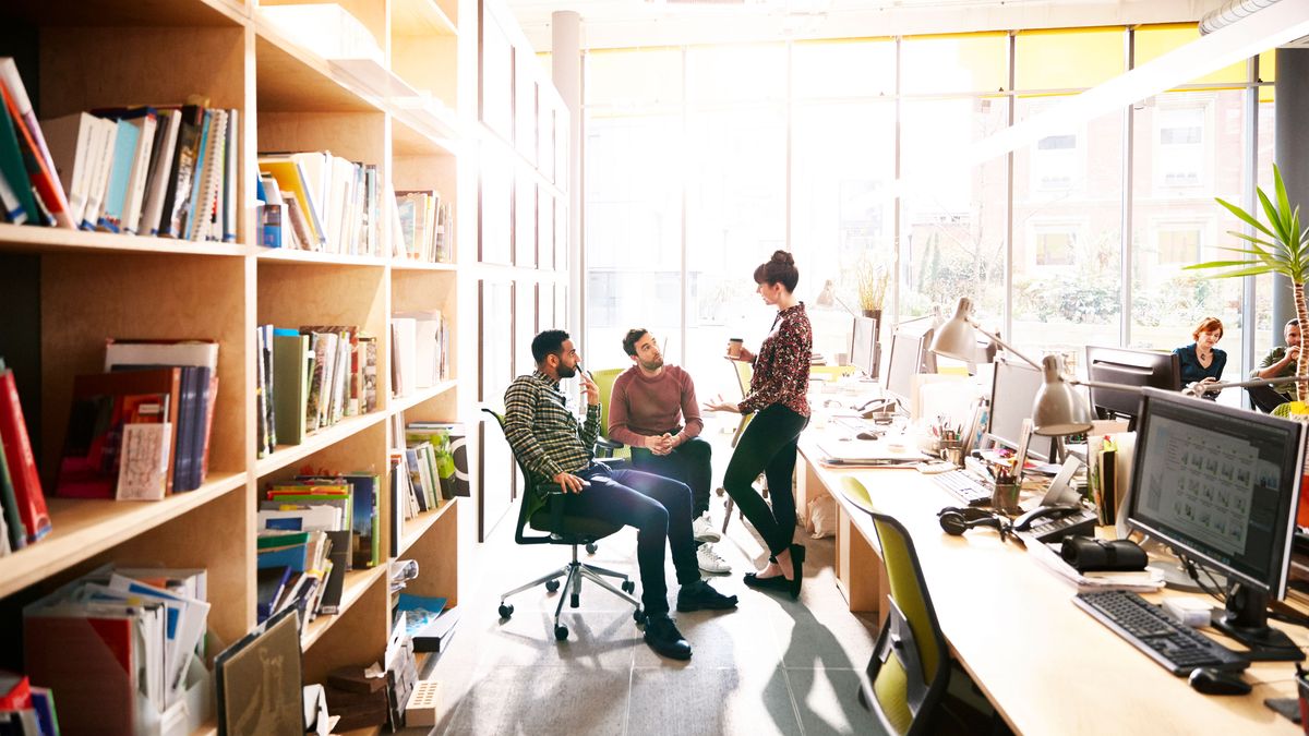 Three colleagues talking in a office between desks with computers and bookshelves