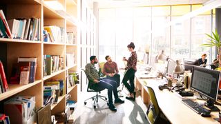 Three colleagues talking in a office between desks with computers and bookshelves